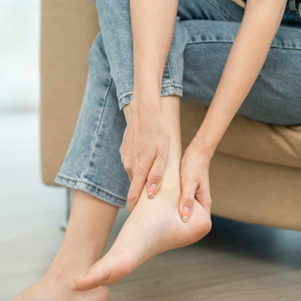 Person sidder på en sofa og holder om sin vrist, hvilket indikerer smerter eller ubehag i foden.