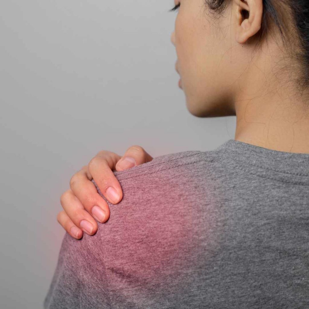 Kvinde med hånden på skulderen som viser tegn på inflammation i skulder med rødme og smerte.