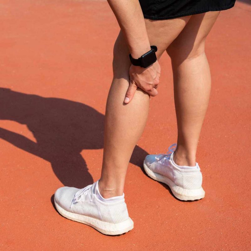 Person i hvide løbesko og sort shorts holder sig om baglåret på en løbebane – illustration af ondt i knæhasen.